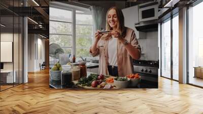 Young woman taking photo of her food with smart phone in modern kitchen at home during isolation and quarantine  Wall mural