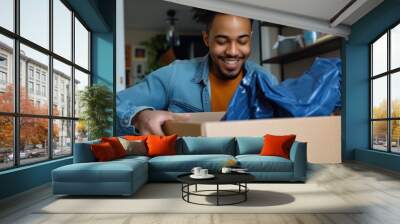 Low angle view of a cheerful multiracial young man at home opening a cardboard box and putting out a cobalt blue running long sleeve top mens.  Wall mural