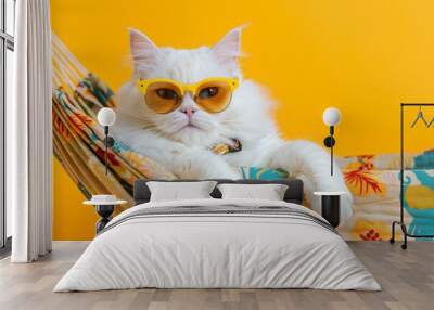 Portrait of an adorable white cat in sunglasses and an shirt, lies on a fabric hammock, isolated on a yellow background. Wall mural