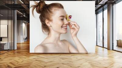 Caucasian woman is covering her eye with a flower posing with bare shoulders on a white studio wall Wall mural