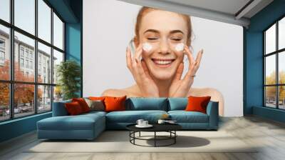 Close up of a beautiful woman with red hair and freckles playing with both hand cream on her face laughing with closed eyes against a white wall. Wall mural