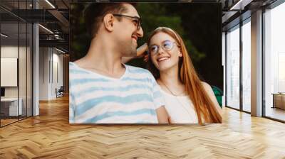 Close up of a charming young couple having fun laughing while sitting on a bench and looking to each other. Wall mural