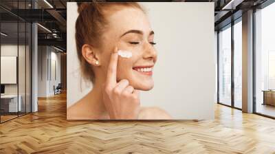 Close up of a young female with red hair and freckles applying white cream on her face laughing with closed eyes isolated on white background. Wall mural