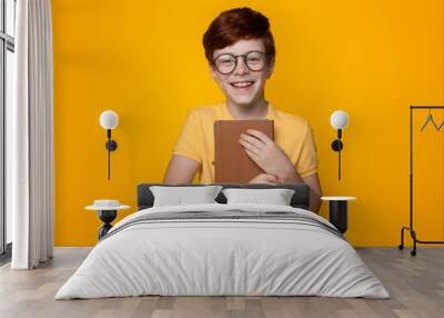 Close up photo of a ginger boy embracing book and wear glasses smiling at camera on a yellow studio wall Wall mural