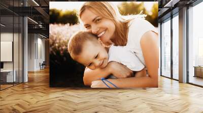 Close up portrait of a lovely young mother embracing his son outside against sunset. Young woman looking at camera laughing while playing with her little kid. Wall mural