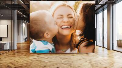 Close up portrait of lovely young mother laughing with closed eyes while her kids is kissing her on the cheeks outdoor against sunset. Wall mural