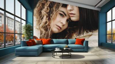 Close up portrait of two serious girlfriend where curly one is posing at photographer while brunette one is embracing her girlfriend and looking away. Wall mural