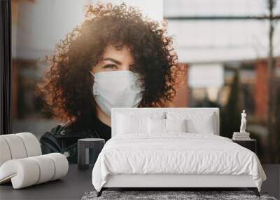 Curly haired caucasian girl is trying to prevent a flu and is wearing a white protective mask Wall mural