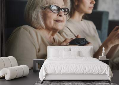 Cute emotional senior woman and daughter playing videogame holding gamepad controllers sitting on sofa at home. Having fun on weekend. Family of two having fun Wall mural