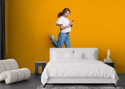 Emotional caucasian blonde woman running and jumping on a yellow studio wall while chatting on mobile and promoting something Wall mural