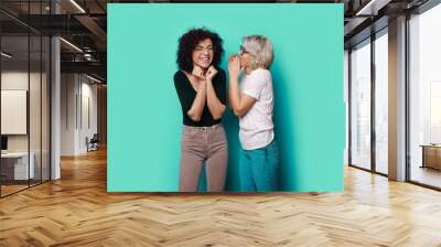 Excited woman listening to friend whispering to her ear on the blue background. Fashion model. Attractive beautiful girl. Wall mural