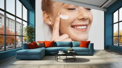 Freckled caucasian ginger lady applying on face anti aging cream and smile on a white studio wall Wall mural