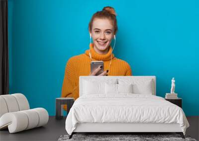 Ginger lady with freckles holding a phone and listening to music while smiling on blue background Wall mural