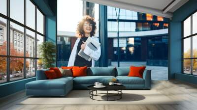 Lovely curly haired entrepreneur with glasses smiling outside while posing with a laptop and tablet Wall mural