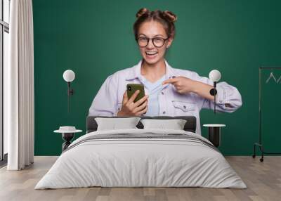 Photo portrait of happy brunette woman pointing finger at holding phone in one hand isolated over green background. Internet communication. Hand holding Wall mural