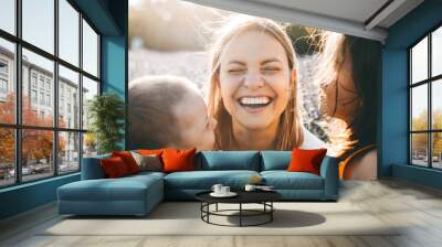 Portrait of a beautiful young mother laughing while closed eyes while her little daughter and son are embracing he in a field of flowers. Wall mural