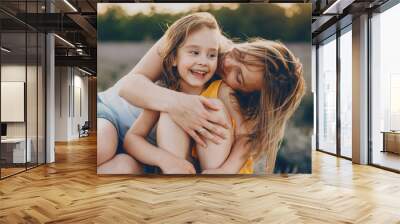 Portrait of a charming young woman embracing her little daughter and kissing her on the cheek while she is laughing at sunset in a field of flowers. Wall mural