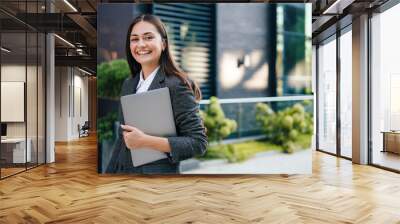 Portrait of a stylish happy business woman with laptop computer in hand walking near office building alone. Outdoor office. Business on the go concept. Wall mural