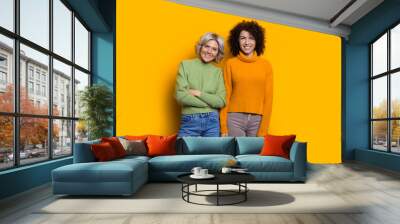 Portrait of two happy lovely sisters one with blonde hair another with curly hair looking at camera laughing isolated on yellow studio wall. Wall mural