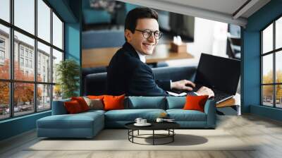 Side view portrait of a stylish adult caucasian manager wearing suit and glasses looking at camera smiling over his shoulder while working at laptop in cafe. Wall mural