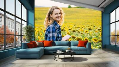 Smiling happy woman farmer working in the field, using a mobile application on a digital tablet in sunflower field. Free sace for text. Concept of smart Wall mural