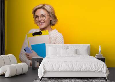 Stunning blonde teacher looking through eyeglasses while holding some books and posing on a yellow wall with freespace Wall mural