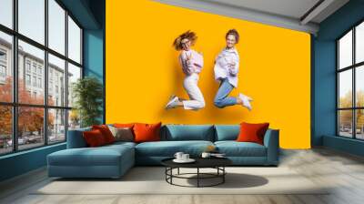 Two blonde sisters jumping on a yellow studio wall and smile at camera gesturing happiness Wall mural