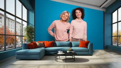 Two friends posing on a blue background. One girl with blond hair, short and straight, but the other with curly and brown hair Wall mural