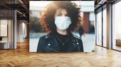 Young curly haired woman wearing a medical mask outside posing at camera in a sunny day Wall mural