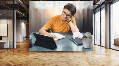 young student with glasses is having online lessons from home using a tablet and writing in a copybook Wall mural