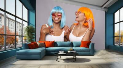 Portrait of two beautiful happy Asian women friends in bright blue and orange wigs and sunglasses posing over gray background Wall mural