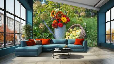 Summer still life. Wicker basket with ripe apples and bunch of red zinnia and sunflowers in vintage watering can on the green lawn Wall mural