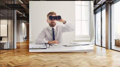 A businessman looks through binoculars while sitting at a table with a computer in the office. Wall mural