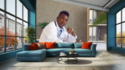 African American black skin doctor in white coat medical professional sitting at table with laptop looking at camera portrait. Modern clinic office interior. Medicine, digital communication technology Wall mural