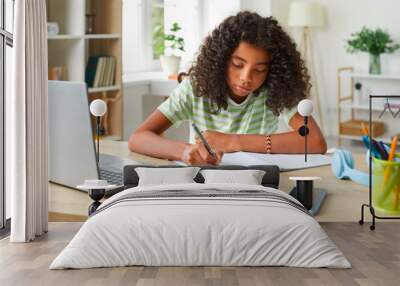 African American teenage girl handwriting in notebook while doing homework using laptop. Concentrated cute curly girl sitting at table in her room and studying remotely. Homeschooling concept. Wall mural