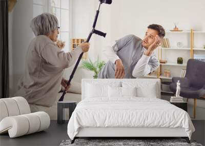 Angry elderly senior woman going to hit young male nurse or doctor with crutch. Enraged exasperated female pensioner standing at home and threatening her scared caretaker with cane. Wall mural