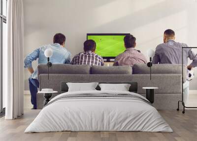 Back view of young men sitting on comfortable grey sofa and watching live football game on TV. Group of 4 friends relaxing on soft couch at home and enjoying interesting soccer match on television Wall mural