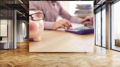 Banner with close up of cute piggy bank wearing eyeglasses placed on table of accountant who's working with tax documentation, analyzing budget and using calculator and laptop computer in background Wall mural