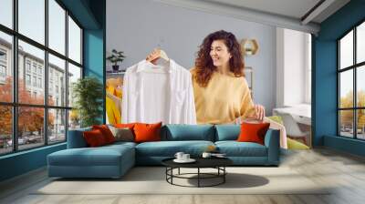 Beautiful woman choosing clothes on rack at home. Attractive smiling young woman holding hangers with shirts, thinking what to wear. Girl trying to choose outfit dressing Wall mural