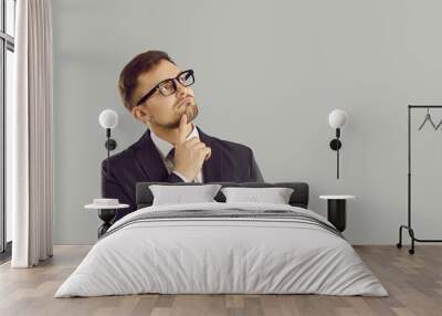 Businessman thinking about something. Young business man in suit and glasses standing isolated on gray background, holding hand on chin and looking at copy space with pensive puzzled face expression Wall mural