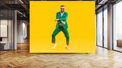 Cheerful and stylish man is having fun dancing in Gangnam style at St. Patrick's Day party. Young man in trendy youth green suit and sunglasses is dancing and laughing on orange background. Banner. Wall mural