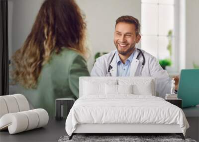 Cheerful friendly male doctor at medical office in hospital, showing examination results on laptop monitor screen and having consultation with young woman patient sitting in clinic. Wall mural