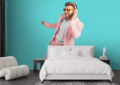 Cheerful happy young man in headphones dancing with happy smile. Joyful excited guy in sunglasses and stylish party suit listening to music and dancing over isolated studio background Wall mural