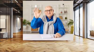 Cheerful senior man having video call, online meeting. Elderly man sitting at table, waving his hand doing hello gesture, looking at camera. Grandfather chatting with friends or family via video call Wall mural