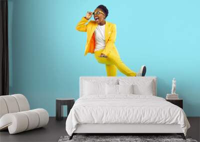 Cheerful young woman having fun, dancing and laughing crazy isolated on light blue background. African American young woman in yellow suit and sunglasses is walking with funny expression on her face. Wall mural