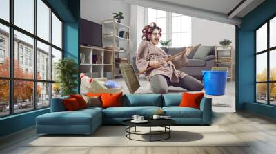 Cheerful young woman having fun and revealing hidden talent while cleaning house. Crazy housewife in hair curlers and face mask tidying up home, singing funny songs and playing on pretend mop guitar Wall mural