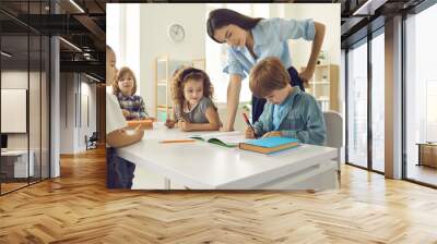 Children learning to write. Happy kids learning new things and acquiring new skills in class. Young school teacher helping her cute little students who are writing in their notebooks Wall mural