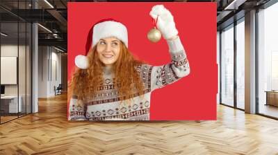 Christmas atmosphere. Joyful young woman in Christmas outfit is looking at Christmas tree ball in her hand. Redhead curly girl in santa hat, christmas sweater and mittens isolated on red background. Wall mural