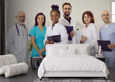 Clinic or hospital staff at work. Diverse team of happy smiling doctors, clinicians, therapists, cardiologists in scrubs and white coat uniform standing together, holding clipboards, looking at camera Wall mural