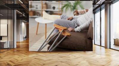 Close up of two crutches leaned on the sofa at home, with a young man who has a plaster cast on his broken leg sitting in the background. Injury concept Wall mural